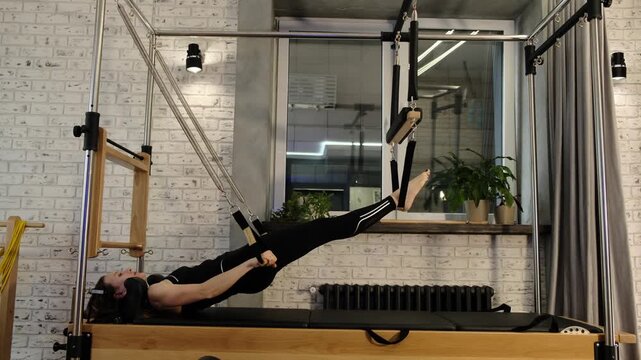 A woman performs an inverted Pilates exercise on a reformer tower, holding straps for balance and control. The frame from workout footage emphasizes strength, focus, and body alignment.