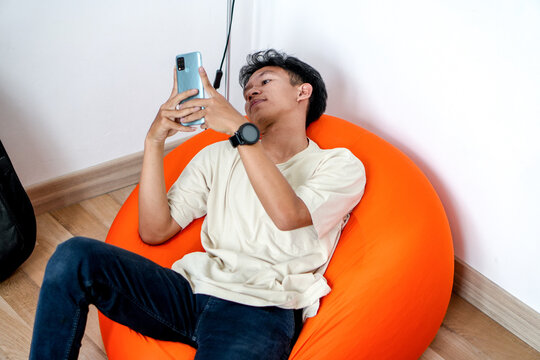 Young Asian Man Relaxing On Beanbag Looking At Phone With Modern Smartwatch