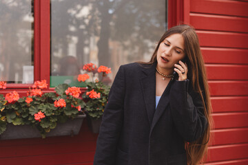 Stylish young woman with brunette long hair and makeup talking on smartphone and standing in trendy outfit, blazer, girl standing near red door background and flowers on window.