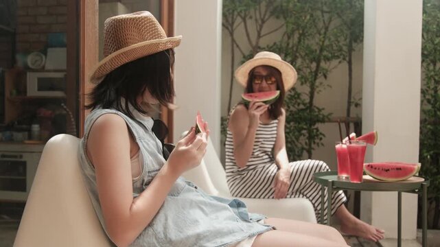 Chilling Asian family holidays, casual girl and mum enjoy eating pieces of fresh red watermelons sliced and smoothie drinks, happiness relaxing on vacation, and hot summertime at outdoor terrace.