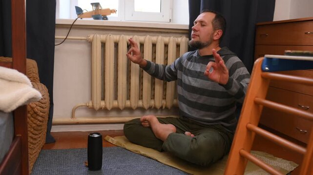 A bearded man listens to music on a portable speaker at home and meditates.
