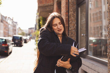 Fototapeta premium Stylish young woman multitasking on a sunny city sidewalk, talking on smartphone while checking a planner. Candid urban portrait conveying productivity, remote work, planning and modern lifestyle.
