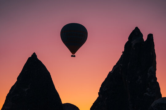 Goreme, Turkiye - 10 October 2019: View of a lone hot air balloon silhouetted against the fiery gradient of the sunset sky, framed by the dark, jagged silhouettes of the fairy chimney rock formations.