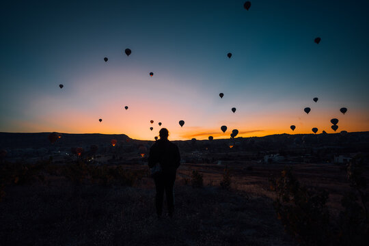 Goreme, Turkiye - 09 October 2019: View of a solitary figure silhouetted against the fiery dawn sky, punctuated by the floating silhouettes of hot air balloons in Cappadocia.
