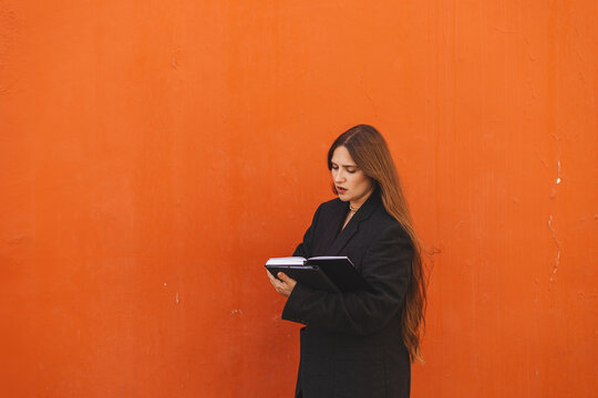 Young woman with long hair reading a notebook, tablet while standing against a bright orange wall, wearing jacket, watch her deals. Minimalist portrait with strong color contrast and natural light.
