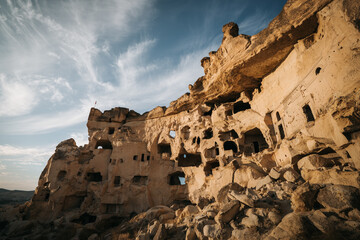 Goreme, Turkiye - 10 October 2019: View of the ancient cave dwellings carved into the soft rock formations, bathed in the warm glow of the setting sun, against a backdrop of a wispy, blue sky.