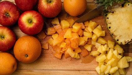 Wooden cutting board holding apples, oranges and diced fruit in kitchen, with pineapple and knife