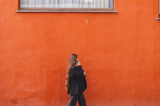 Serious young adult business woman walks and talks on phone, solves problems, holds documents, notebook, tablet and bag on background of orange wall. Girl dressed in black jacket, denim jeans. Side.