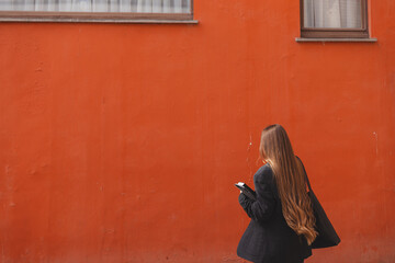 Naklejka premium Serious young adult business woman walks and talks on phone, solves problems, holds documents, notebook, tablet and bag on background of orange wall. Girl dressed in black jacket, denim jeans. Back.