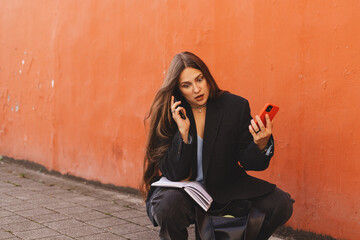 Young business woman outdoors multitasking with two phones, surprised and busy while working remotely. Concept of communication overload, modern lifestyle, digital stress and entrepreneurship.