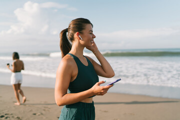 Smiling athletic woman using wireless earphones and phone during morning routine, embracing digital tools to stay motivated and connected while moving.
