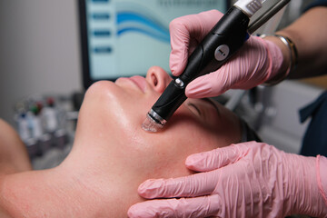 Woman receiving professional hydrafacial with a handheld device from an aesthetician in pink gloves, modern clinic setting focused on cleansing, exfoliating and hydrating skin for a healthy glow