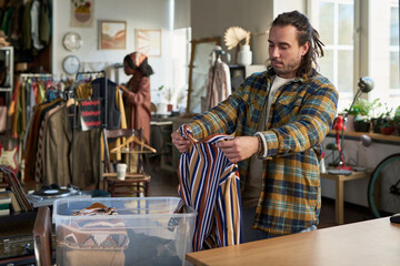 Caucasian young adult man sorting striped shirt in thrift shop, standing near plastic bin with folded clothes while Black young adult woman browsing clothing racks in background