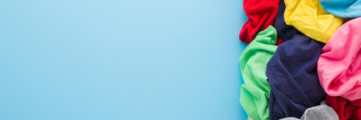 Heap of colorful t shirts on light blue table background. Pastel color. Different messy clothes. Closeup. Wide banner. Empty place for text. Top down view.