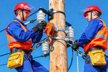 An electrician wearing seat belts, helmet and gloves works on high-voltage power lines and equipment attached to a wooden pole against a clear blue sky
