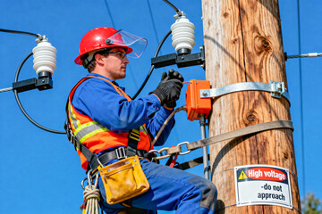 An electrician wearing seat belts, helmet and gloves works on high-voltage power lines and equipment attached to a wooden pole against a clear blue sky