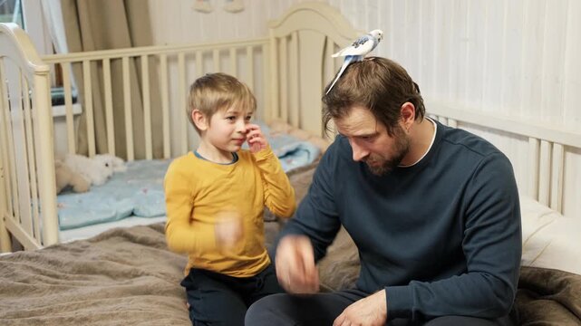 Father and son enjoying at home. Young boy and his daddy playing with funny pet budgerigar parrot. Happy family. Tamed budgie parakeet. People take care of and play with bird. Cute domestic animals