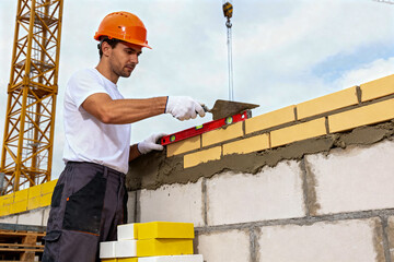 Professional Bricklayer or Construction Worker Wearing an Orange Hard Hat and Gloves, Using a Trowel and Level to Precisely Lay Yellow Bricks on a Construction Site with a Crane in the Background