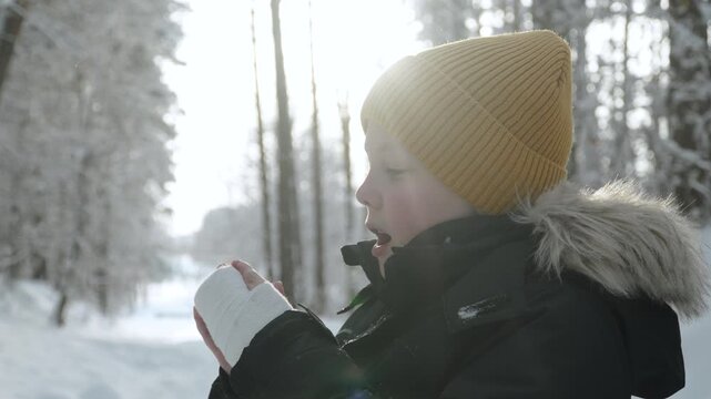 Child tries to warm hands with hot breath in forest during freezing winter weather. Side view portrait of boy in warm clothing and cast on arm, warming himself on snowy day.