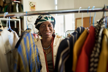 Portrait of young adult Black woman smiling while browsing clothing rack in thrift shop, wearing patterned headscarf and necklace, surrounded by assorted garments on hangers