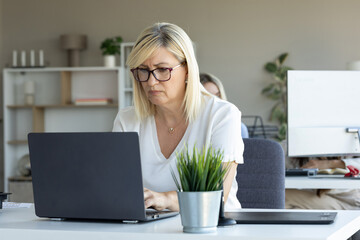 A diligent blonde businesswoman works intently on her laptop in a bright, modern office setting
