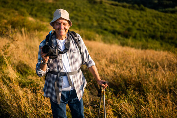 Portrait of happy and active senior hiker using camera for photographing while hiking with backpack and hiking poles in nature.	