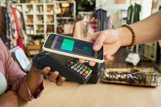 Caucasian young adult man holding smartphone over payment terminal making contactless payment, while Black woman holding card reader in thrift shop with clothing and accessories in background - Powered by Adobe