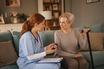 Obraz premium Young female healthcare worker showing medicine bottle to senior woman at her home. The elderly patient is sitting on the sofa holding a cane while listening.