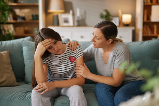 A teenage girl expresses frustration while seated on a sofa, clutching her forehead. Her mother sits beside her, gently encouraging and comforting her in a warm living room atmosphere.
