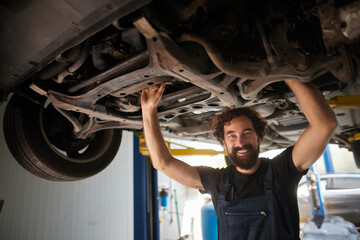 A cheerful mechanic works underneath a lifted car at a busy repair shop. He inspects the vehicle while using tools, ensuring everything is in order for the client.