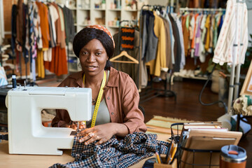 Portrait of young adult Black woman operating sewing machine, working on garment repair at thrift shop, measuring tape around neck, clothing racks and shelves visible in background