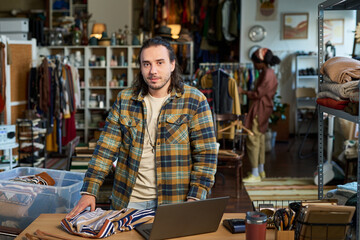 Fototapeta premium Portrait of adult Caucasian man standing behind counter folding clothes in thrift shop, looking at camera with neutral expression, laptop and folded garments on table, shelves and racks in background