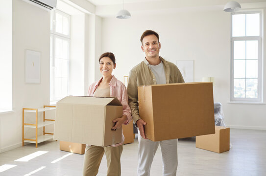 Happy couple moving cardboard boxes into new apartment. Sunlit living room with big windows, minimal furniture and scattered packing gear. Relocation, new beginnings at home and real estate
