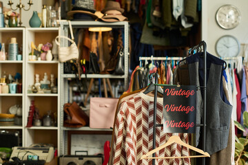 Clothing rack displaying assorted vintage garments standing in foreground of thrift shop with shelves and racks filled with accessories, bags, and hats visible in background