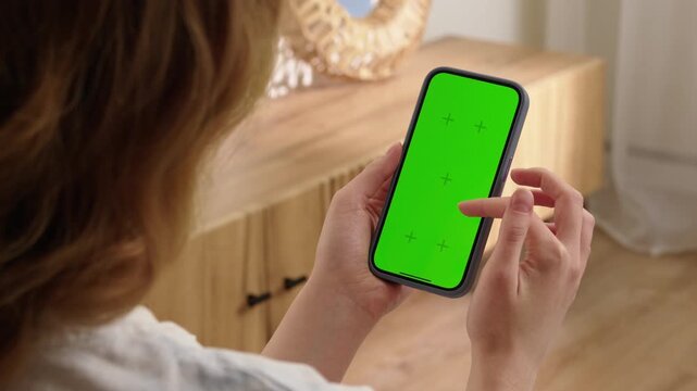 Woman sit use digital tech cell phone with green screen mock up. Hand scroll phone web site, swipe like photos. Girl hold mobile closeup. Chroma key page. Cozy home background. Online app copy space.