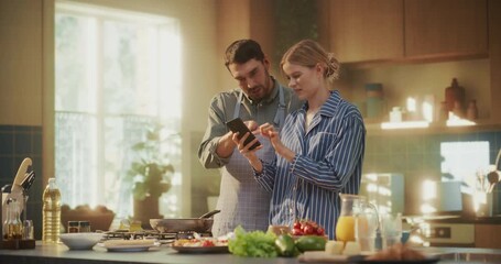 Relaxed Morning Routine in a Cozy Apartment Kitchen. Couple Cooking Breakfast, Flipping Food in a Frying Pan and Checking Smartphone for Instructions and Inspiration Online - Powered by Adobe