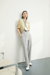 Fashionable woman in beige blouse and gray trousers standing in a modern indoor setting.