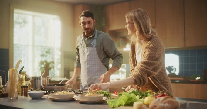 Happy Couple in a Bright Kitchen Sharing Dinner Moments. Man Serves Pasta on a Plate while His Girlfriend Makes the Final Touches and Adding Herbs, Both Enjoying a Peaceful Evening Together