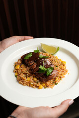 Hands hold a white plate with sliced, seared duck breast drizzled with a dark sauce, resting on fried rice with corn and cilantro, garnished with a lime wedge