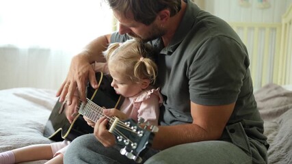 Happy dad teaches his cute daughter to play the guitar while sitting on the sofa in the living room at home. Happy Father's Day. Father teaching his child to play guitar. Kid Learning music skill - Powered by Adobe