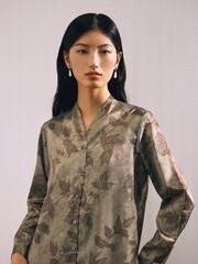 Elegant woman in floral patterned blouse, standing in relaxed pose, conveying modern warmth in a simple indoor setting.