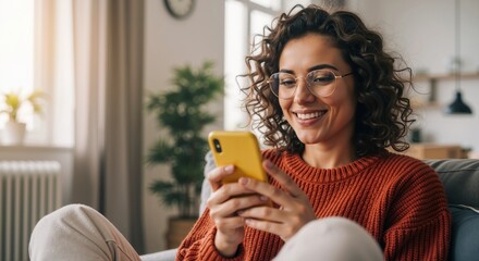 Woman with curly hair and glasses smiling while looking at and using a yellow smartphone, relaxing on a sofa in a bright modern home living room. Digital communication with copy space