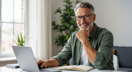 Senior Caucasian man with grey hair and glasses smiling at camera while working on laptop in modern home office. Remote work and successful business lifestyle, banner with copy space