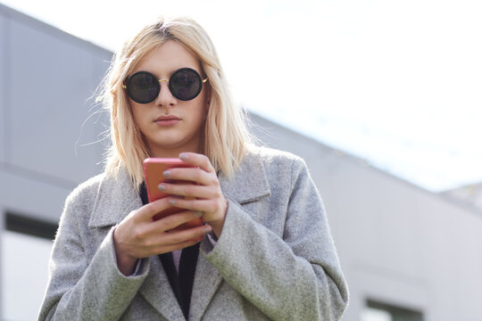 Portrait of fashionable transwoman. Low angle view of blond-haired transgender in round sunglasses and stylish coat holding smartphone and texting message