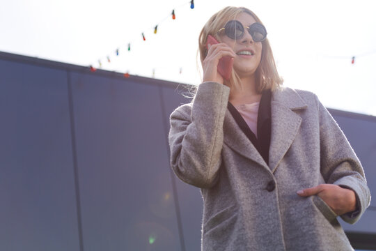 Portrait of androgynous businesswoman. Low angle view of confident stylish blond transwoman in sunglasses talking on smartphone and smiling
