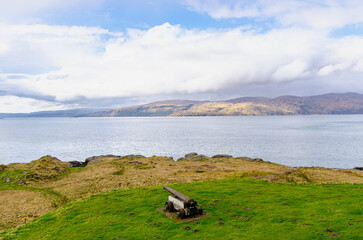 Historic cannon pointing toward the sea on grassy terrain, overlooking a tranquil bay surrounded by distant hills and cloudy sky.