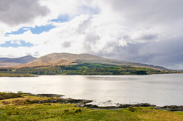 Beautiful view of a calm lake with rolling hills and mountains in the background under soft sunlight and dramatic clouds.