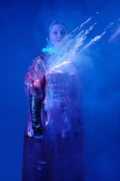 Vertical medium long studio portrait of senior woman wearing futuristic metallic outfit with corrugated tubes around neck standing in navy blue light with steam out of chest effect, gen Z fashion inte
