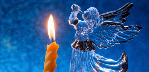 Glass Christmas angel blowing a trumpet beside a lit spiral candle on glittering blue bokeh; herald...
