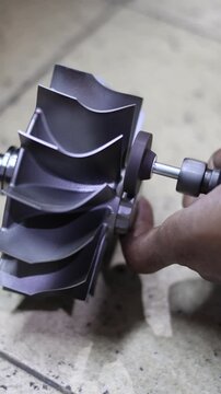 Close-up of a worker's hand using a bor tuner or borcun to perform a correction or balance adjustment on the metal blades of a turbocharger compressor wheel.
